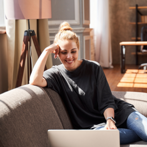 Mulher branca de cabelo preso, blusa escura, sentada no sofá, sorrindo em frente ao computador, fazendo psicoterapia online.